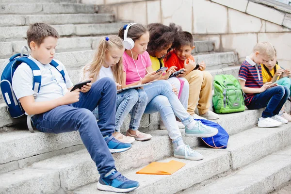 Group of schoolchildren of different nationalities, in colorful clothes sitting on stone steps. Teenagers talking, listening to music on headphones, reading books.