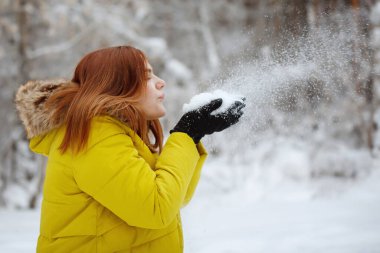 Genç, güzel, kızıl saçlı, hardal ceketli uzun saçlı kadın kışın karlı bir ormanda ellerinden kar taneleri salıyor..