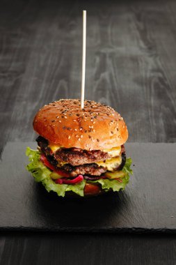 Düz siyah tabakta ev yapımı burger. Susamlı çörek, peynirli köfte, marul, soğan, kürdanla dikilmiş domates..