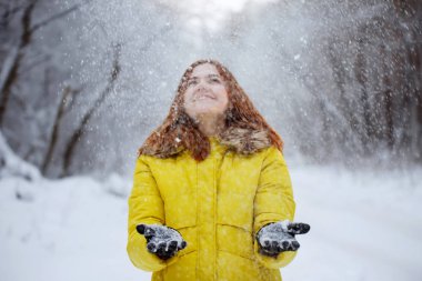 Genç, güzel, kızıl saçlı, hardal ceketli uzun saçlı kadın kışın karlı bir ormanda ellerinden kar taneleri fırlatıyor..