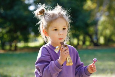 Mor takım elbiseli küçük kız yazın şehir parkında çikolata yiyor.