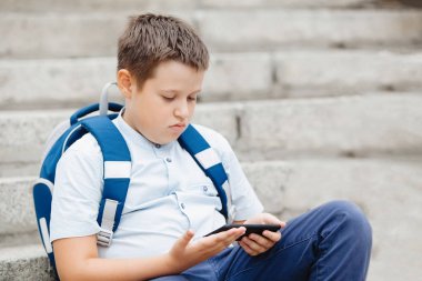 Sırt çantalı çocuk açık havada güneşli bir yaz gününde merdivenlerde oturan akıllı telefona bakıyor..