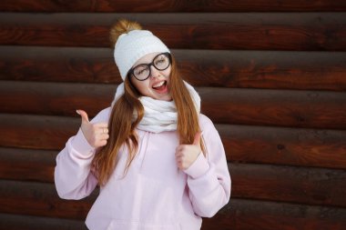 Genç, gözlüklü, beyaz örgü şapkalı ve kazaklı güzel bir kadın, başparmağının ahşap kahverengi bir tahta duvarın arka planında olduğunu gösteriyor. Gençlik, sağlıklı yaşam tarzı, hayaller.