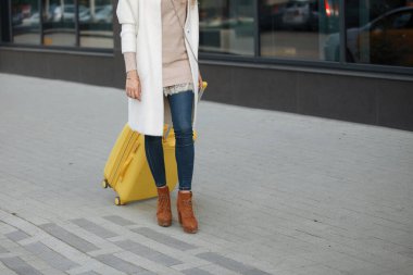 Otelin yakınında yürüyen bej ceketli, sarı valizli, ayak bileği çizmeli bir kadın. Seyahat konsepti. Yakın plan..