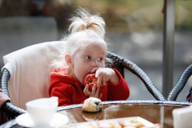 Little blonde girl with a tail eats an apple at a table in a cafe.