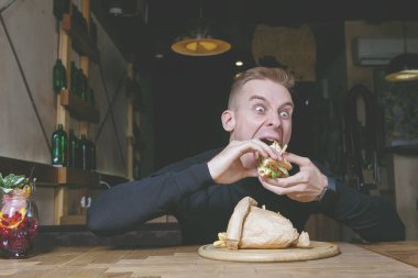 Sarışın bir adam kafedeki bir masada sandviçi ısırıyor..