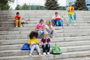 Farklı milletlerden bir grup öğrenci, renkli giysiler içinde taş basamaklarda oturuyorlar. Gençler konuşuyor, kulaklıkla müzik dinliyor, kitap okuyor..