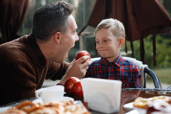 Father and son are sitting at a table in a cafe. A man offers a boy to eat an apple. Parental love, happy childhood, child care.