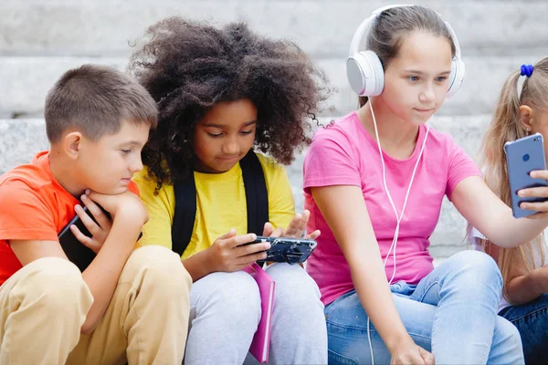Group of schoolchildren of different nationalities, in colorful clothes sitting on stone steps. Teenagers talking, listening to music on headphones, reading books.