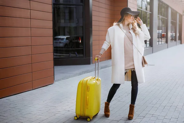 Woman in ankle boots in a hat with a yellow suitcase in a beige coat walking near the hotel. Travel concept.
