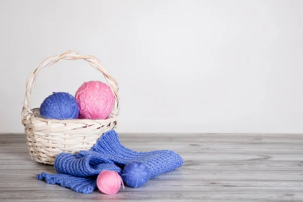 Basket with balls of wool and hand-knitted scarves in blue and pink. Gray background.