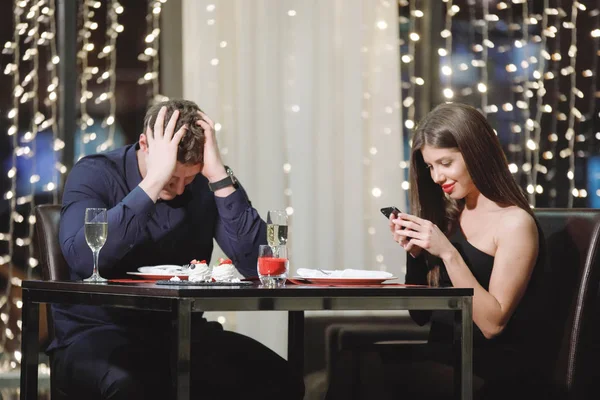 Conflict couple at a table in a restaurant. Woman looking at smartphone. Man is bored, clasping his hands in his head