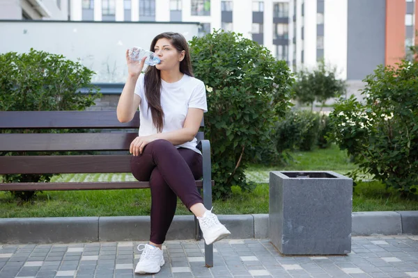 Woman drinks water from a plastic bottle, sits on a bench in an urban environment in the summer.