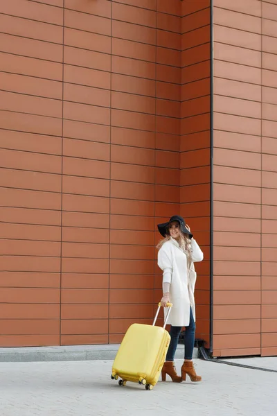 穿着脚踝靴 头戴一顶黄色行李箱 身穿米黄色外套的女人走在酒店附近 旅行概念 — 图库照片