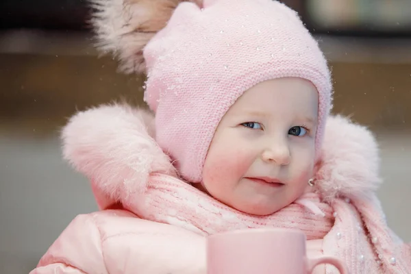 ピンクの女の子の赤ちゃんは ポンポンのウィンクでキャップの全体をゆっくりとウィンクします 都会の公園での晴れた冬の日 — ストック写真