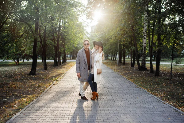 Loving couple hugging on a pavement alley in the park. He is in a gray coat and black glasses, she is in white and in a hat.
