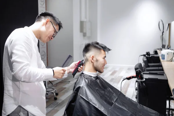 Young guy a hairdresser cuts a hair clipper on a man\'s head in a beauty salon.