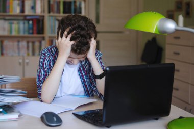 Genç bir çocuk elinde dizüstü bilgisayarla bir masaya oturur ve kafasını eline tutuşturur. Evde koronavirüs karantinası eğitimi alıyorum. Seçici odaklanma. Bulanık arkaplan.