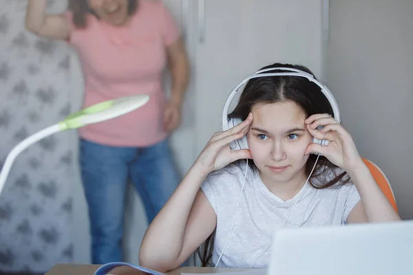 在带笔记本电脑的桌旁 一个戴耳机的少女在教育上的困难 模糊的母亲在后面咒骂 科罗纳威斯隔离了家庭教育 有选择的重点 模糊的背景 — 图库照片