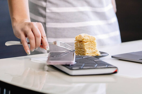 Женщина держит слоеный торт в тарелке на столе. Размытый дневник чашки кофе, ноутбука и смартфона. Избранный фокус. Домашнее задание по карантину коронавируса, социальная дистанция, частный предприниматель.