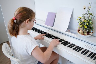 Gözlüklü ve beyaz tişörtlü genç kız oturur, piyano çalar. Evde eğitim, sosyal mesafe. Karantina koronavirüsü. Seçici odaklanma. Bulanık arkaplan.