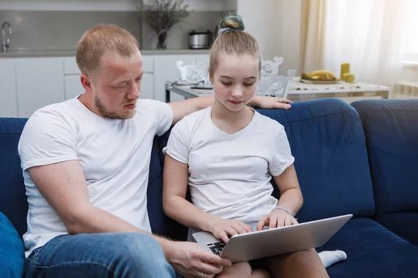 Father and daughter teenager in white t-shirts are sitting on the sofa with a laptop. Joint homework classes. Homeliness. Selective focus. Blur background. Old film filter.