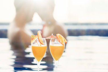 Havuzdaki bir çiftin arka planında dilimler olan iki bardak portakal suyu. Bir kadın ve bir erkek mavi suda yüzüyorlar. Seçici odaklanma. Bulanık arkaplan.