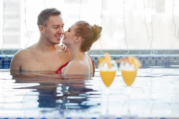 两杯橙汁和切片放在游泳池里的一对夫妇的背景上 一个女人和一个男人在蓝色的水里游泳 有选择的重点 模糊的背景 — 图库照片