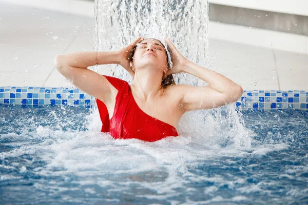 穿着红色游泳衣的金发女人躺在游泳池的水流下 蓝色的水滴在酒店的温泉里 — 图库照片