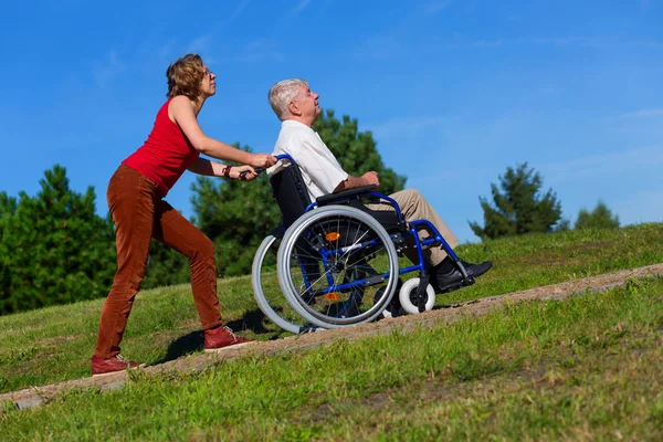 Młoda kobieta wcisnąć stary człowiek na wózku inwalidzkim do góry hil — Zdjęcie stockowe