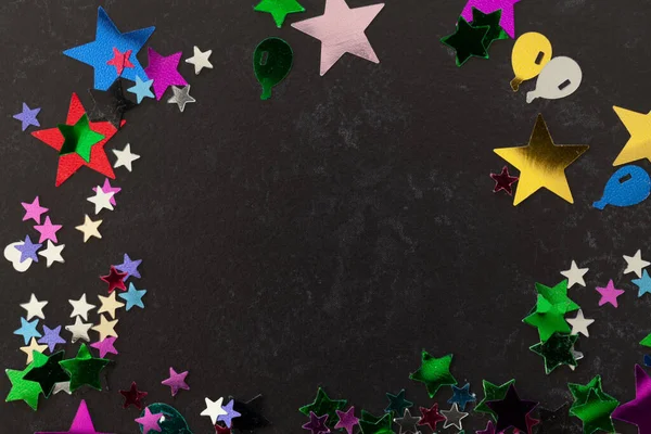 Party confetti stars and shaped of all colours scattered on a black table with copy space