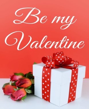 Beyaz, kare hediye kutusu kırmızı arka plan beyaz masada kırmızı ve beyaz puantiyeli kurdele ile süslenmiş. İpek kırmızı gül yanındaki kutuyu döşeme. Sevgililer mesaj eklendi