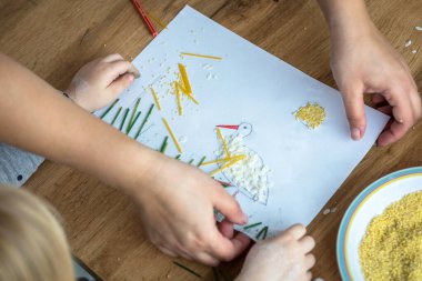 Anaokulu öğrencisi evde makarna ve kuskus kullanarak resim yapıyor.
