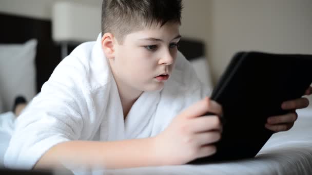 Adolescente en albornoz blanco en la cama y jugando en la tableta — Vídeos de Stock