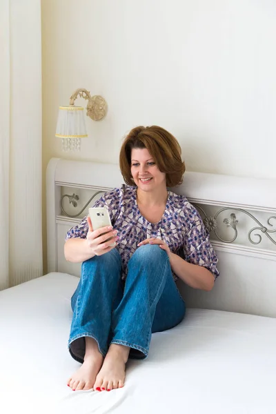 Wanita menikmati smartphone duduk di tempat tidur di kamar tidur — Stok Foto
