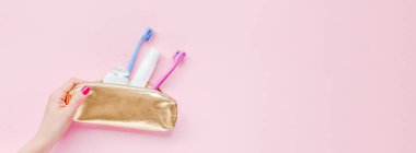 Altın seyahat kozmetik çantasında diş hijyeni ve diş bakımı ürünleri. Pastel pembe arka planda. Kadının elinde manikürlü fotokopi alanı var. Düz konum, üst görünüm bileşimi