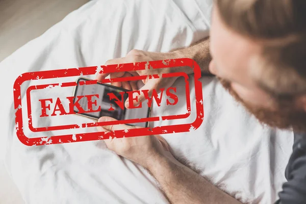 Fake news infodemics during Covid-19 pandemic concept. Man hands holding mobile phone reading global news or social net at home. Reading last news update in lockdown. Red text fake news on foreground