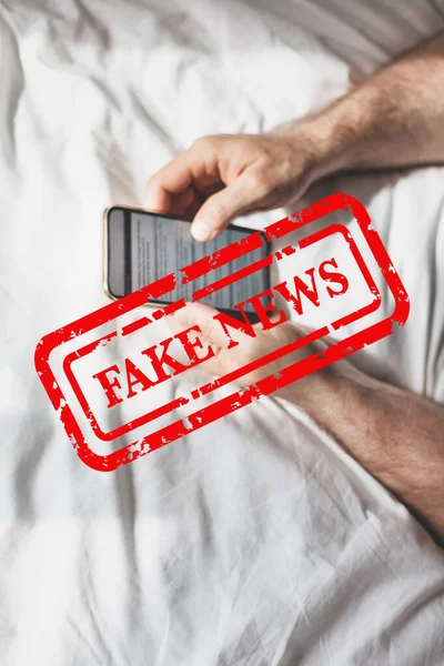 Fake news infodemics during Covid-19 pandemic concept. Man hands holding mobile phone reading global news or social net at home. Reading last news update in lockdown. Red text fake news on foreground