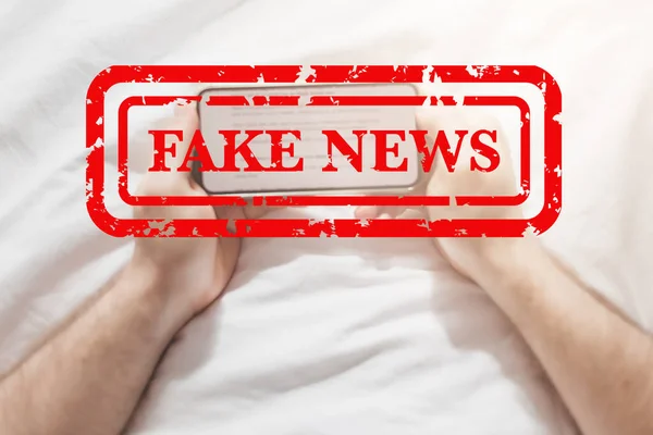 Fake news infodemics during Covid-19 pandemic concept. Man hands holding mobile phone reading global news or social net at home. Reading last news update in lockdown. Red text fake news on foreground