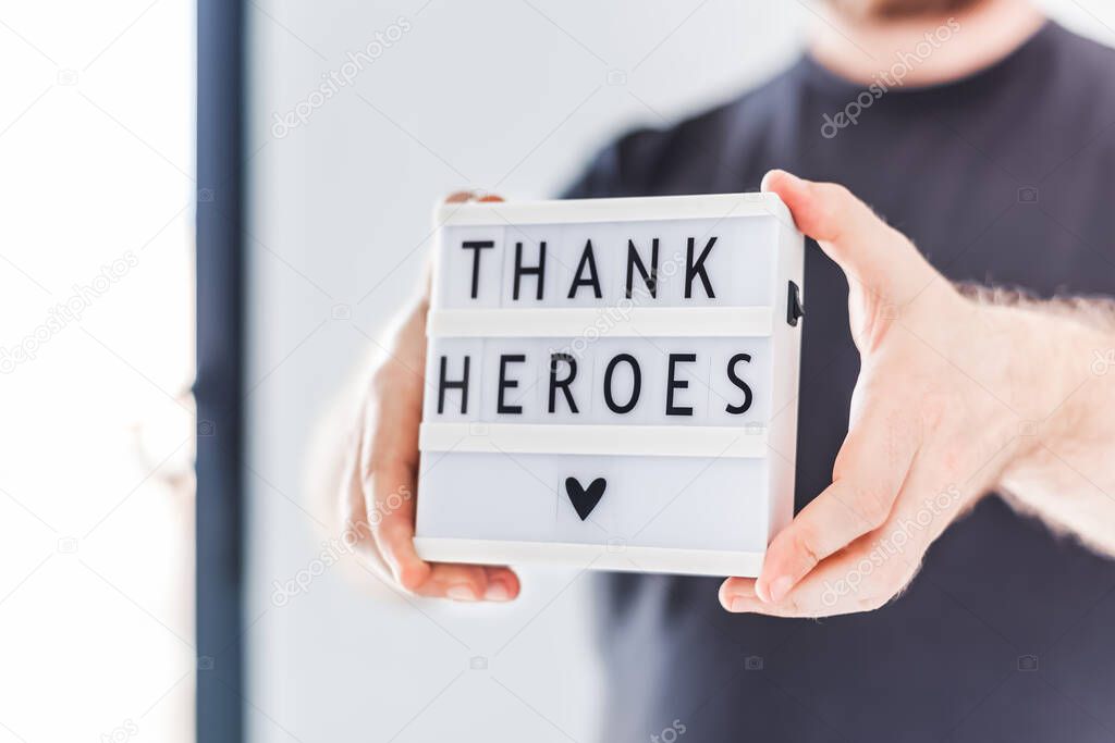 Nurse day concept. Man hands holding lightbox with Thank heroes text thanking doctors, nurses and medical staff working in hospitals during coronavirus COVID-19 pandemics. View through window glass