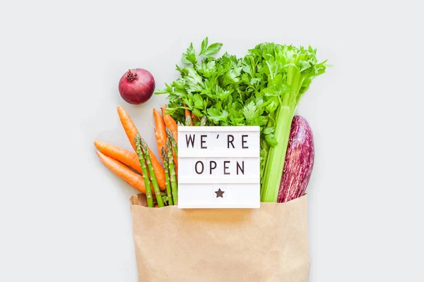 End of quarantine. Fresh organic vegetables in eco craft paper shopping bag with text We\'re Open lightbox flat lay, top view on gray background. Sustainable lifestyle. Zero waste, plastic free concept
