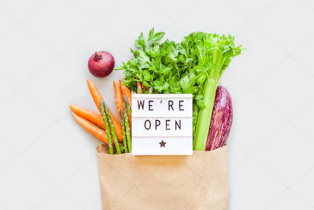 End of quarantine. Fresh organic vegetables in eco craft paper shopping bag with text We're Open lightbox flat lay, top view on gray background. Sustainable lifestyle. Zero waste, plastic free concept