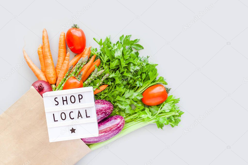 Fresh organic vegetables in eco craft paper shopping bag with text Shop Local on lightbox flat lay top view with copy space on gray background. Sustainable lifestyle. Zero waste, plastic free concept.