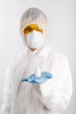 Tek kullanımlık anti-salgın takım elbiseli doktor solunum cihazı takıyor ve gözlük beyaz arka planda haplarla dolu bir şişe tutuyor.