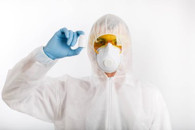 Tek kullanımlık anti-salgın takım elbiseli doktor solunum cihazı takıyor ve gözlük beyaz arka planda haplarla dolu bir şişe tutuyor.