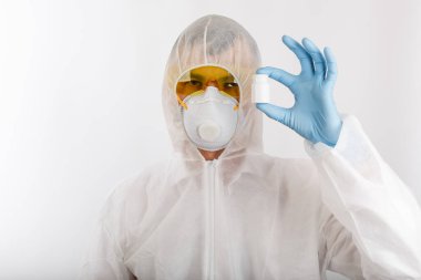 Tek kullanımlık anti-salgın takım elbiseli doktor solunum cihazı takıyor ve gözlük beyaz arka planda haplarla dolu bir şişe tutuyor.