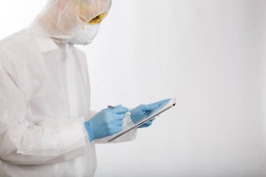 Salgın önleme giysisi ve solunum cihazı giymiş bir doktor beyaz arka planda kişisel elektronik tablet ve metin yeri üzerine notlar alıyor.