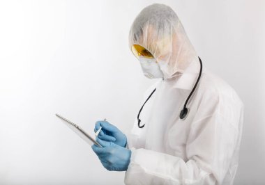 Salgın önleme giysisi ve solunum cihazı giymiş bir doktor beyaz arka planda kişisel elektronik tablet ve metin yeri üzerine notlar alıyor.