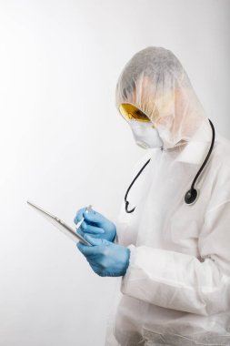 Salgın önleme giysisi ve solunum cihazı giymiş bir doktor beyaz arka planda kişisel elektronik tablet üzerine notlar alıyor.