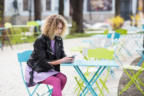 黒革のジャケットに巻き毛の長い美しい女性の肖像画と紫のドレスは スマートフォンで公園のカフェテーブルに座って — ストック写真